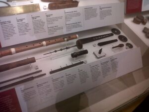 photograph of a museum display case with parts of different musical instruments, including pipes, laid out with descriptor cards