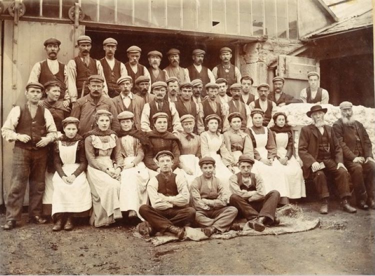 Old image of 4 rows of workers, male and female, wearing working clothes - men in flat caps and women with aprons