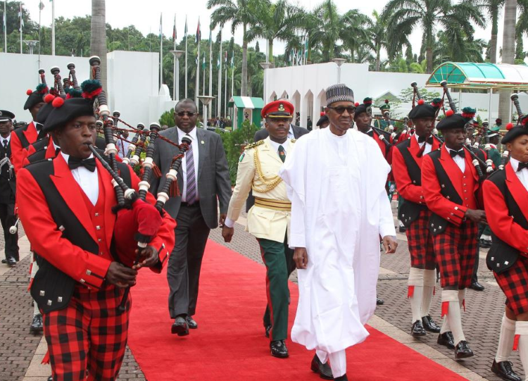 A red carpet, with an important man wearing white robes walking down the middle, flanked by a double row of bagpipers wearing tartan trousers and military dress jackets and hats.