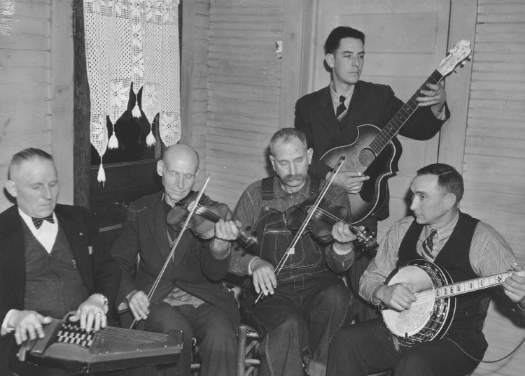 A folk band of 5 players, 4 seated (dulcimer, banjo and fiddles) and one standing up (guitar)