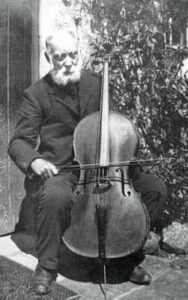 man seated outside a cottage door, playing a cello one-handed