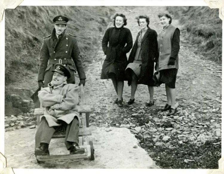 3 women laughing at a soldier pushing another soldier in a wheelbarrow